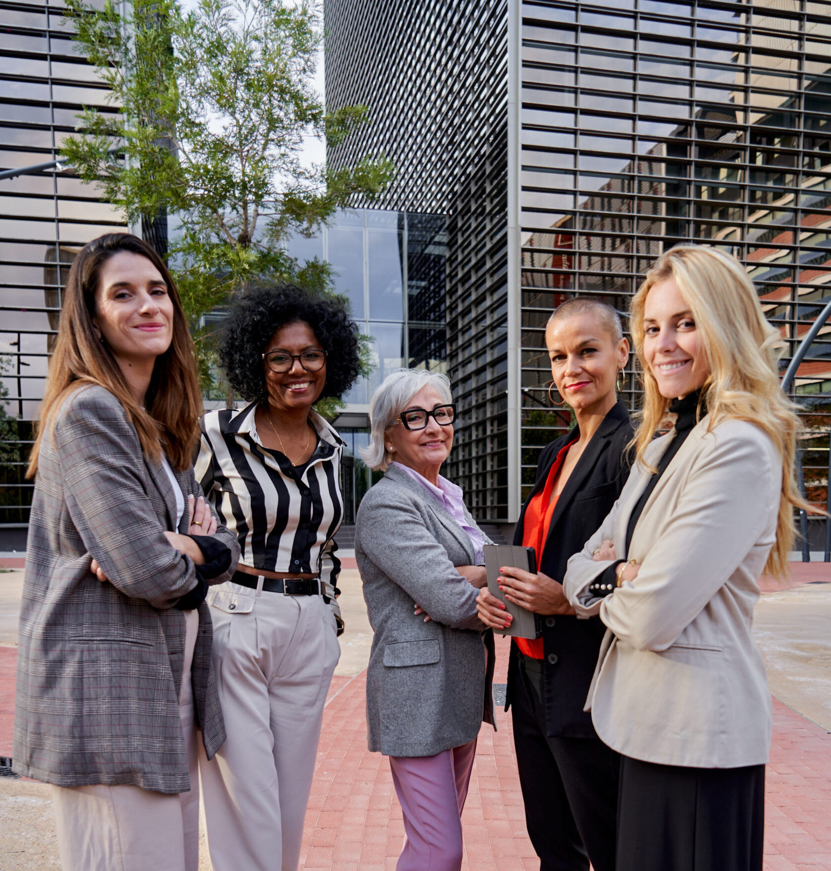 Cheerful group of multiethnic businesswomen looking at camera smiling, headed by a mature woman, standing outside financial district. Vertical photo.